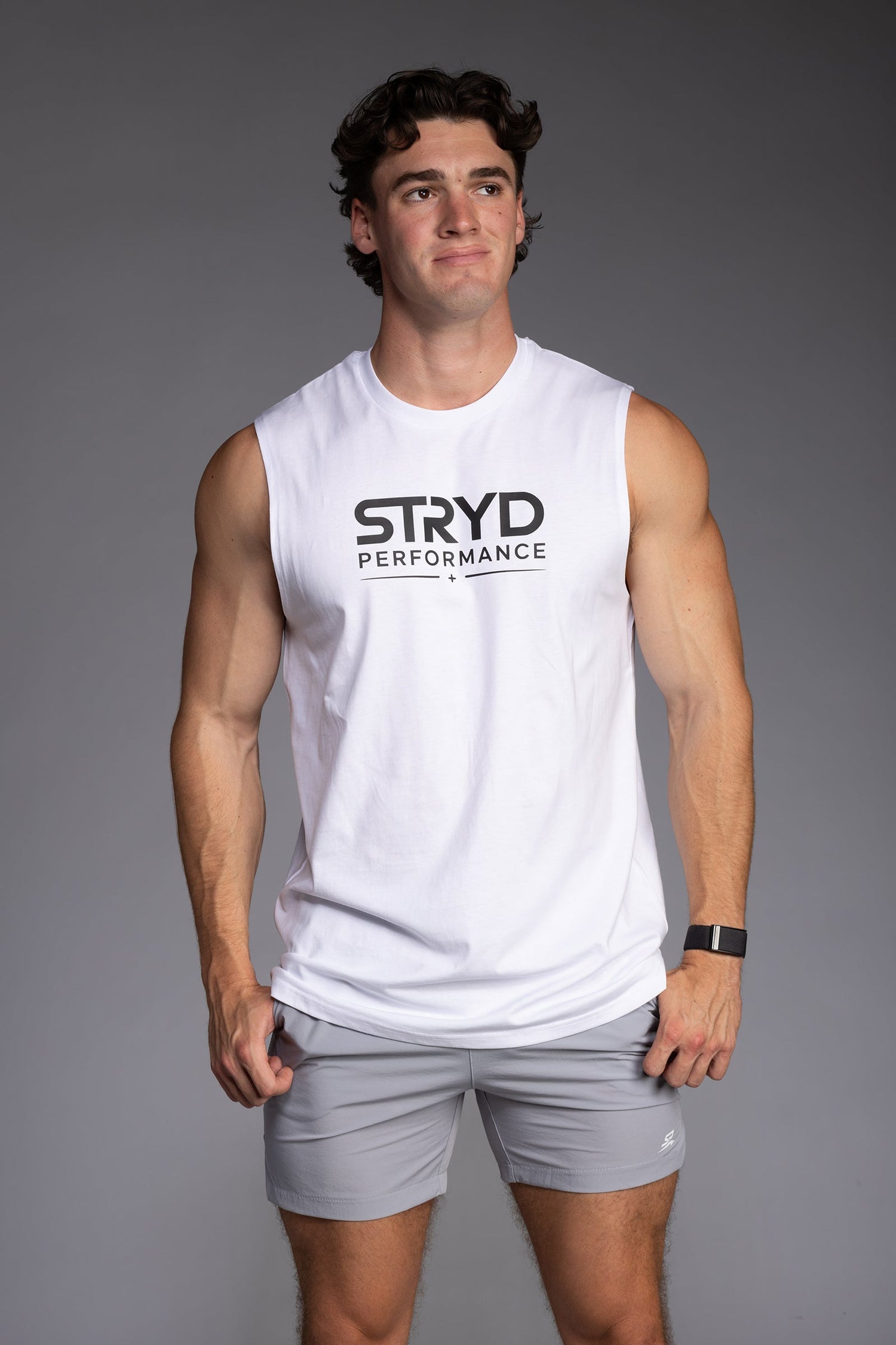 muscular male model wears white Premium Tank top with Light Grey training shorts against grey wall - STRYD Performance
