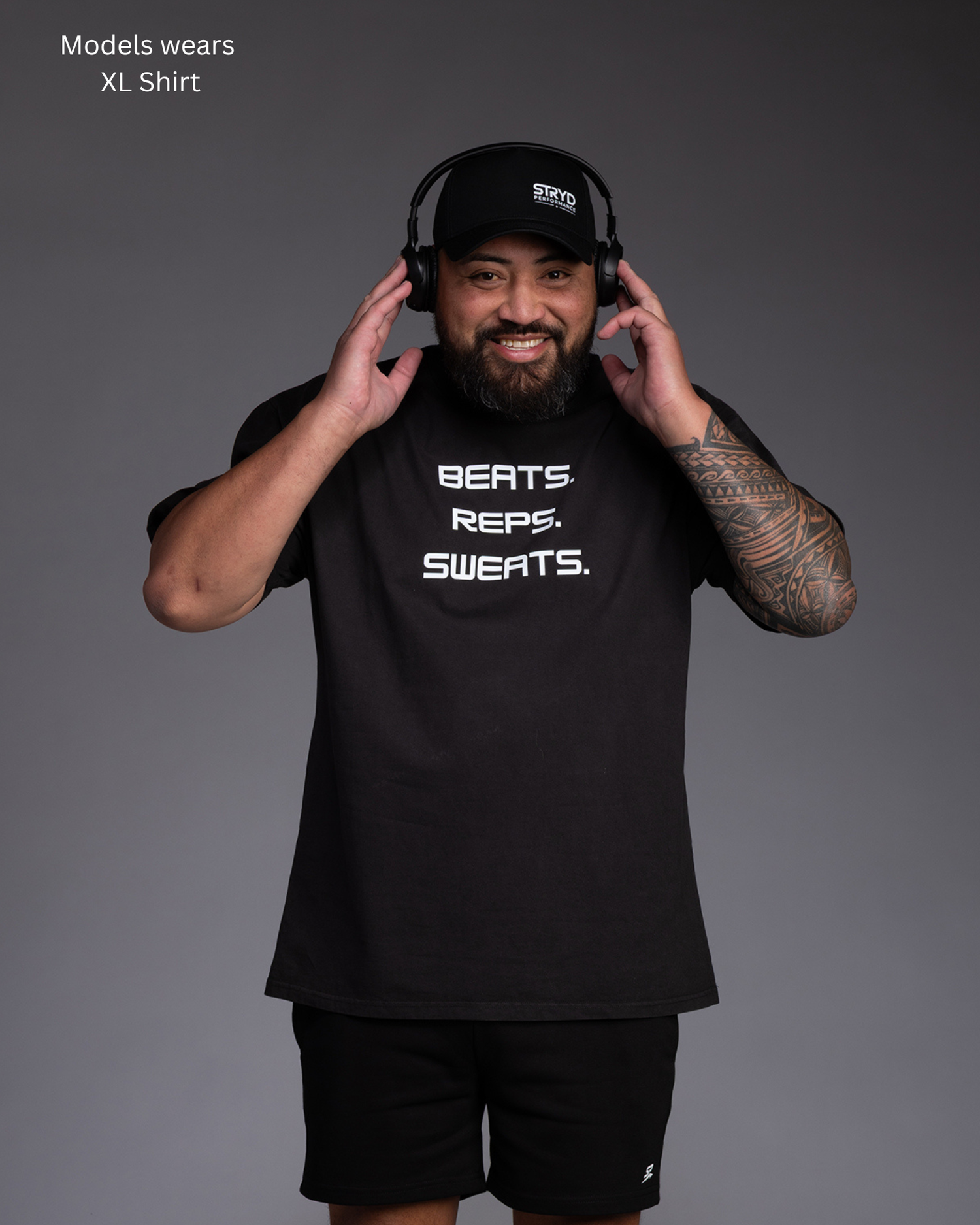 Man wearing an oversize faded Black shirt with the words Beats Reps Sweats on the front in White lettering holding his headphones to his ears and listening to tunes - STRYD Performance I like it loose shirt