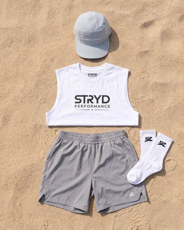 Flat lay on sand: run cap, white tank, grey training shorts and crew socks